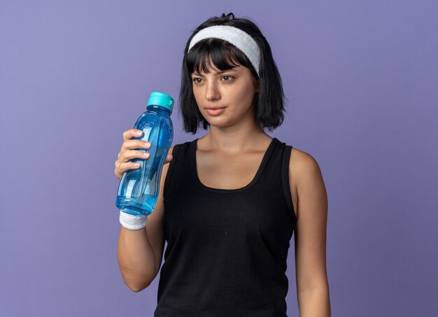 青い背景の上に立っている深刻な顔で脇を見て水筒を保持しているヘッドバンドを身に着けている若いフィットネスの女の子