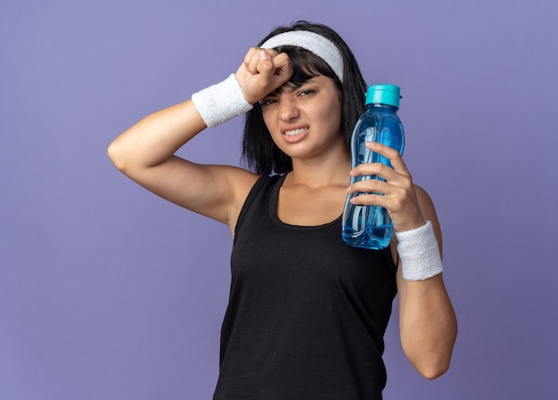 Young fitness girl wearing headband holding water bottle looking tired and overworked standing over blue background