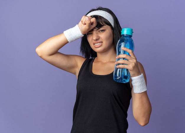 Молодая фитнес-девушка с повязкой на голову, держащая бутылку с водой, выглядит уставшей и перегруженной, стоя на синем фоне
