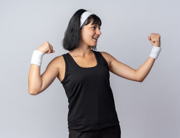 Young fitness girl wearing headband looking aside happy and cheerful raising fists standing over white