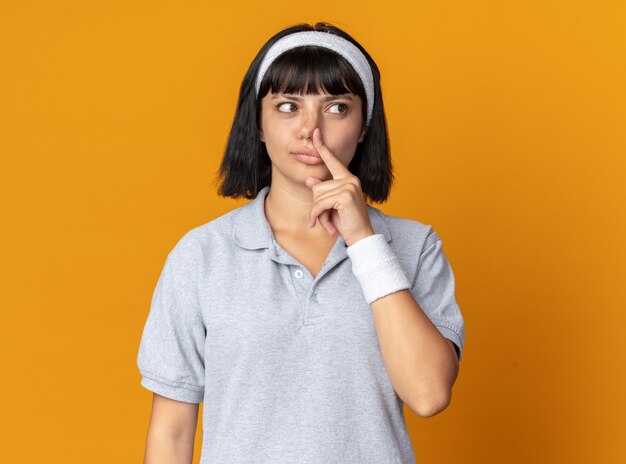 Young fitness girl wearing headband looking aside puzzled closing her nose with finger 