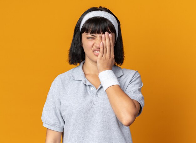 Young fitness girl wearing headband looking at camera confused and displeased covering eye with hand standing over orange