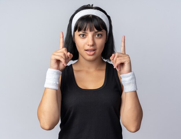 Молодая фитнес-девушка в повязке на голову, смотрящая в камеру, счастлива и удивлена, показывая указательные пальцы, стоящие над белой
