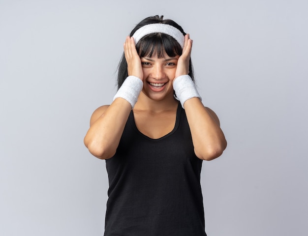 Young fitness girl wearing headband looking at camera hapy and cheerful smiling standing over white