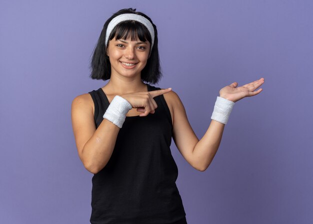 Free photo young fitness girl wearing headband looking at camera presenting copy space with arm of hand pointing with index finger to the side smiling confident standing over blue background