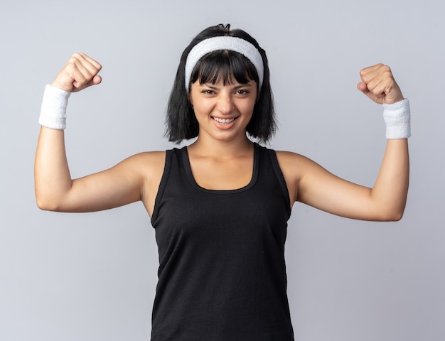 Young fitness girl wearing headband looking at camera strained and confident raising fists standing over white background