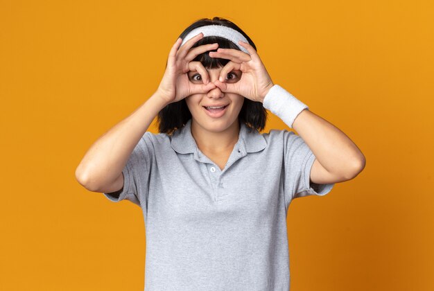 Молодая фитнес-девушка с повязкой на голову смотрит в камеру через пальцы, делая бинокулярный жест, весело улыбаясь, стоя на оранжевом фоне