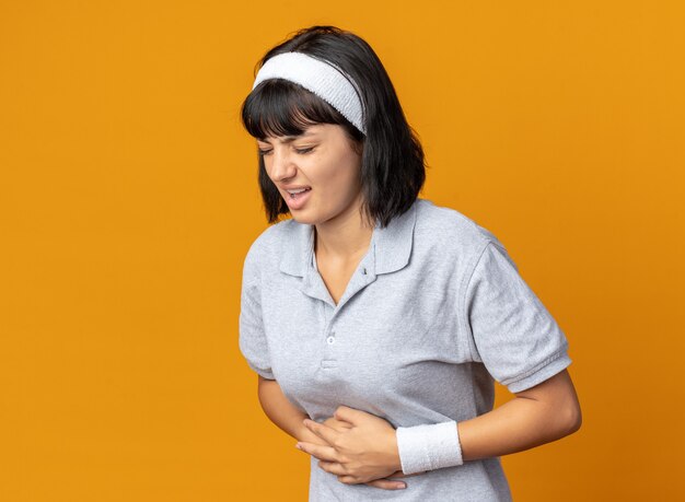 Young fitness girl wearing headband looking unwell touching her belly feeling pain standing over orange background