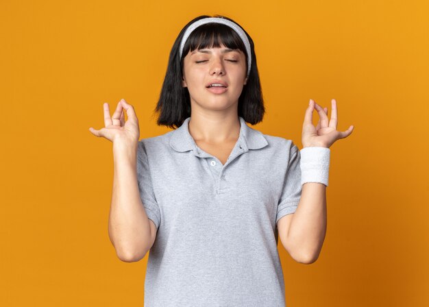 Young fitness girl wearing headband making meditation gesture with fingers relaxing with eyes closed standing over orange