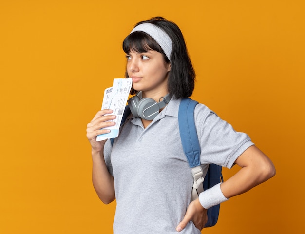 Young fitness girl wearing headband with headphones around neck holding air tickets looking aside puzzled standing over orange background