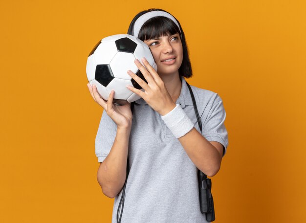 Young fitness girl wearing headband with skipping rope around neck holding soccer ball looking aside displeased standing over orange background