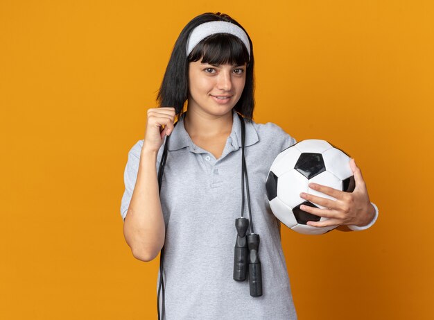 Young fitness girl wearing headband with skipping rope around neck holding soccer ball looking at camera smiling confident raising fist standing over orange background