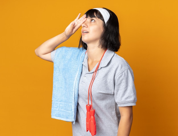 Young fitness girl wearing headband with skipping rope around neck and towel on a shoulder looking tired and bored with hand on her forehead standing over orange background