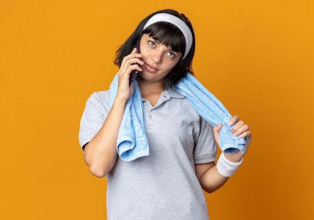 Young fitness girl wearing headband with towel around her smiling confident while talking on mobile phone standing over orange background