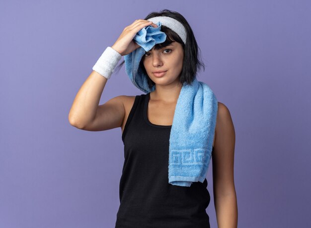 Young fitness girl wearing headband with towel on shoulder wiping her forehead looking tired standing over blue background