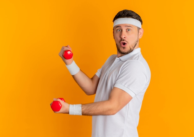 Молодой фитнес-мужчина в белой рубашке с повязкой на голову тренируется с гантелями, глядя вперед, улыбаясь, стоя над оранжевой стеной
