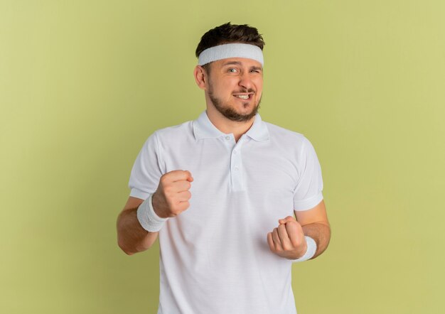 Young fitness man in white shirt with headband, clenching fists looking confused standing over olive wall