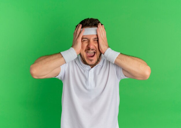 Young fitness man in white shirt with headband crazu happy screaming for success standing over green background