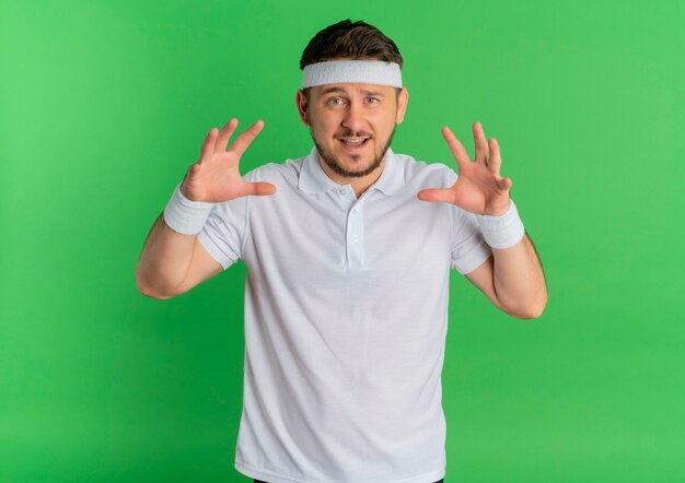 Young fitness man in white shirt with headband doing claw gesture as cat, smiling standing over green background