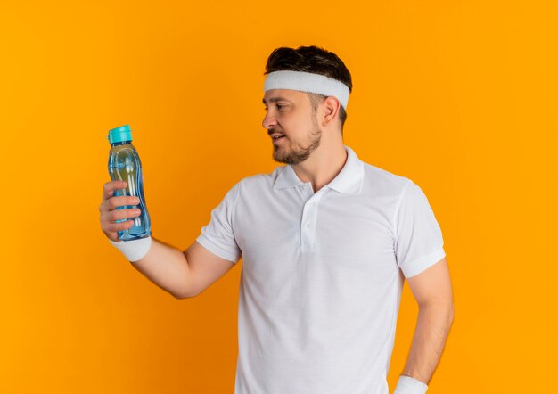 Young fitness man in white shirt with headband holding bottle of water looking at it standing over orange background