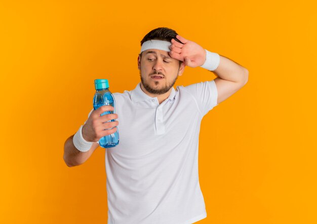 Young fitness man in white shirt with headband holding bottle of water looking tired and exhausted standing over orange background