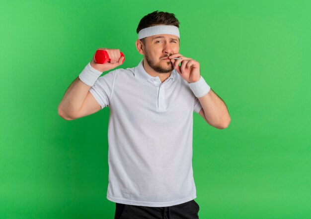 Молодой фитнес-человек в белой рубашке с повязкой на голову, держащий гантели, делает упражнения и курит сигарету, спортивная концепция плохой привычки, стоящая на зеленом фоне