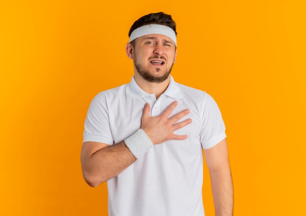 Молодой фитнес-мужчина в белой рубашке с повязкой на голову, держащий руку на груди, уставший и измученный, глядя в камеру, стоит на оранжевом фоне