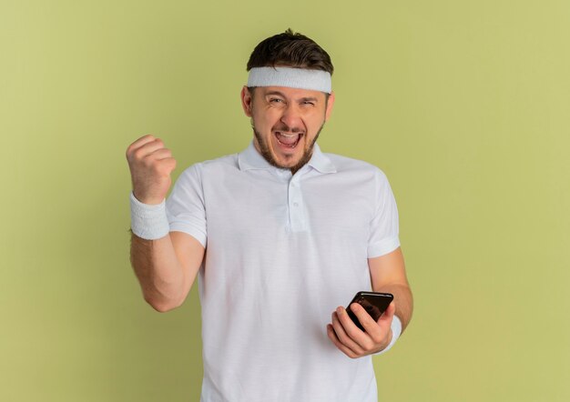 Young fitness man in white shirt with headband holding smartphone raising fist happy and excited standing over olive background