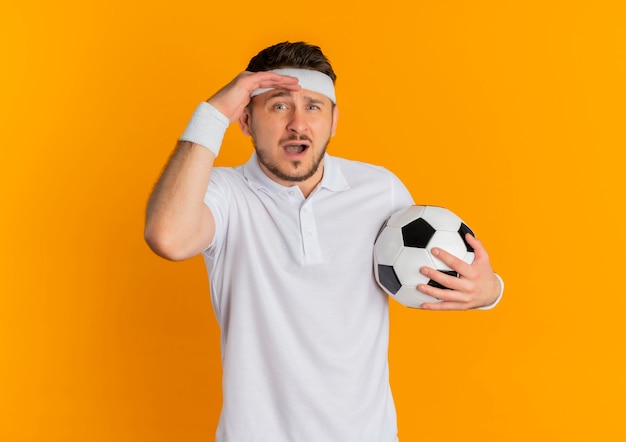 Young fitness man in white shirt with headband holding soccer ball looking at camera confused standing over orange background