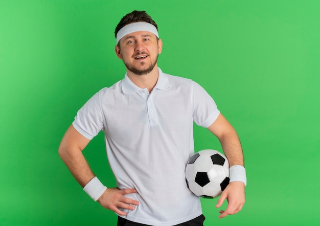 녹색 배경 위에 유쾌하게 서있는 카메라를보고 축구 공을 들고 머리띠와 흰 셔츠에 젊은 피트 니스 남자