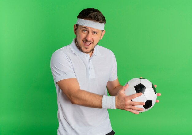 Молодой фитнес-мужчина в белой рубашке с повязкой на голову держит футбольный мяч, глядя в камеру, весело улыбаясь, стоя на зеленом фоне