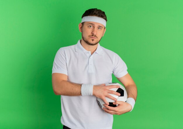 녹색 배경 위에 서있는 자신감이 식으로 카메라를보고 축구 공을 들고 머리띠와 흰 셔츠에 젊은 피트 니스 남자