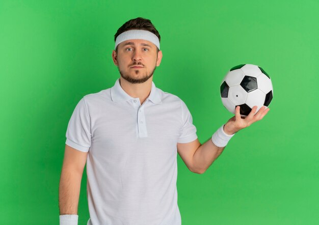 녹색 배경 위에 서 심각한 얼굴로 카메라를보고 축구 공을 들고 머리띠와 흰 셔츠에 젊은 피트 니스 남자