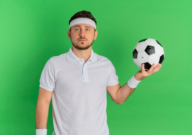 Молодой фитнес-мужчина в белой рубашке с повязкой на голову держит футбольный мяч, глядя в камеру с серьезным лицом, стоящим на зеленом фоне