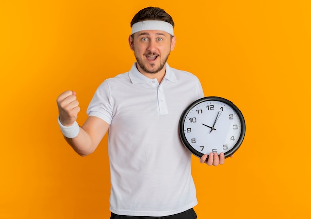 オレンジ色の背景の上に立って幸せで興奮した拳を握り締める壁時計を保持しているヘッドバンドと白いシャツの若いフィットネス男