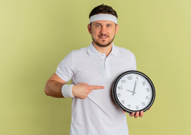 Young fitness man in white shirt with headband holding wall clock pointing with finger to it smiling confident standing over olive background