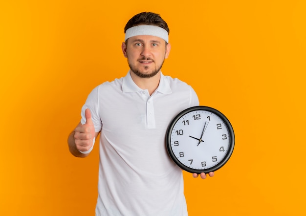 Молодой фитнес-мужчина в белой рубашке с повязкой на голове, держа настенные часы, показывает палец вверх, уверенно стоит на оранжевом фоне