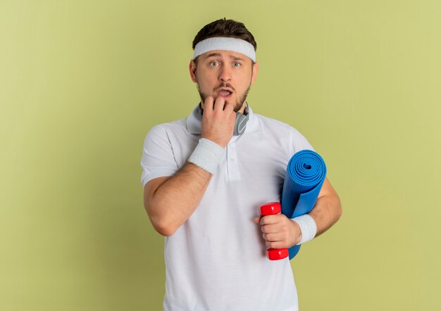 Young fitness man in white shirt with headband holding yoga mat and dumbbell looking at camera surprised standing over olive background