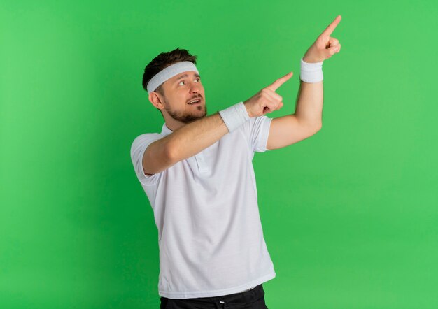 Young fitness man in white shirt with headband looking aside pointing with index fingers to the side standing over green background