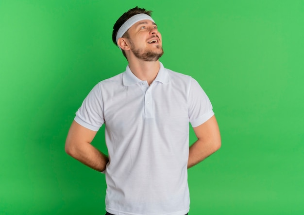 Young fitness man in white shirt with headband looking aside with smile on face standing over green background