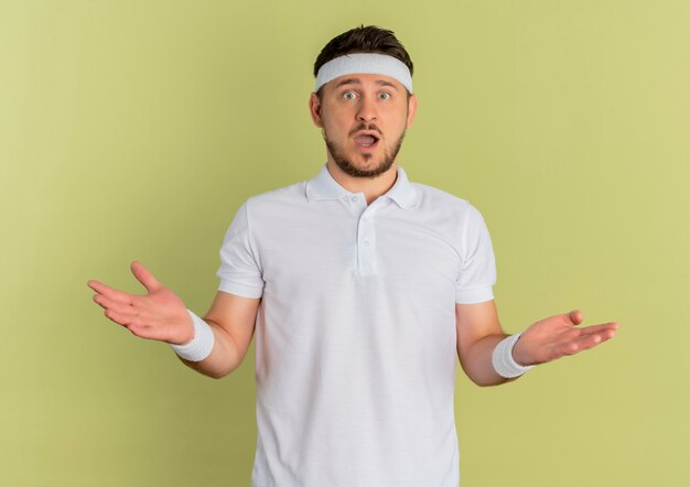 Young fitness man in white shirt with headband looking at camera confused and surprised standing over olive background