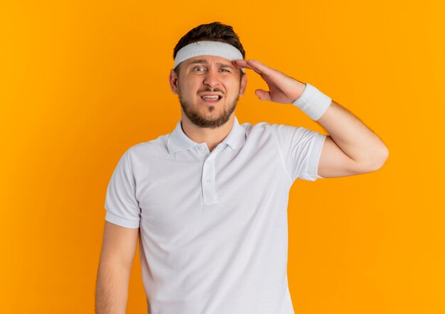 オレンジ色の壁の上に立っている頭の上の手と混同して正面を向いているヘッドバンドと白いシャツを着た若いフィットネス男