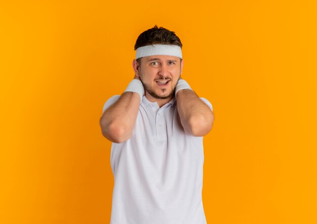 Young fitness man in white shirt with headband looking to the front feeling discomfort standing over orange wall