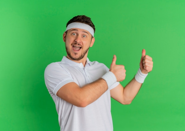 Young fitness man in white shirt with headband looking to the front smiling cheerfully showing thumbs up standing over green wall