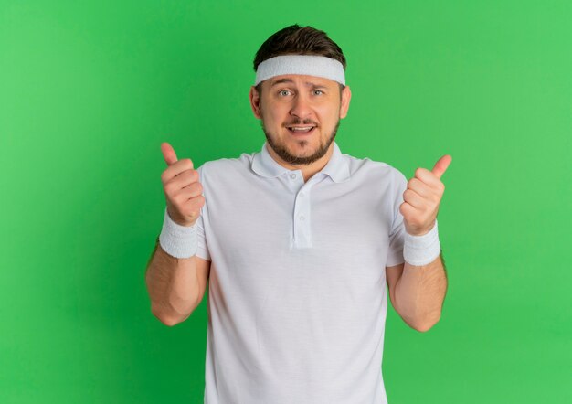 Young fitness man in white shirt with headband looking to the front smiling showing thumbs up standing over green wall