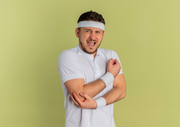 Young fitness man in white shirt with headband looking to the front touching his elbow having pain standing over olive wall