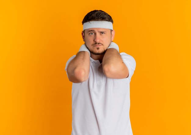 Young fitness man in white shirt with headband looking to the front with sad expression on face standing over orange wall