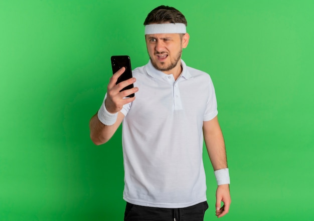 Young fitness man in white shirt with headband looking at screen of his mobile with confused expression standing over green wall