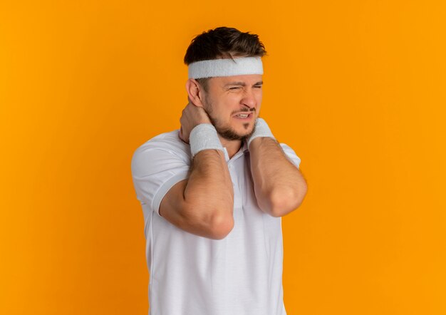 Молодой фитнес-мужчина в белой рубашке с повязкой на голове выглядит нездоровым, касаясь его шеи, чувствуя боль, стоя над оранжевой стеной