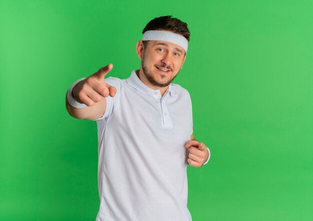 Young fitness man in white shirt with headband pointing with finger to camera with smile on face standing over green background